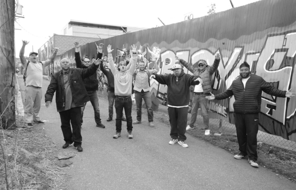 A group of men in front of a wall with graffiti, arms wide, smiling at the camera.