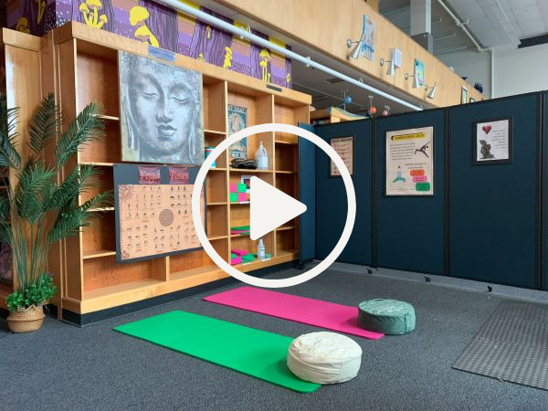 A picture with a play button superimposed over it: Decompression space with shelves that have yoga supplies, a poster of yoga poses, and a close up painting of a restful buddha face. The space includes two yoga mats with meditation pillows on the floor .
